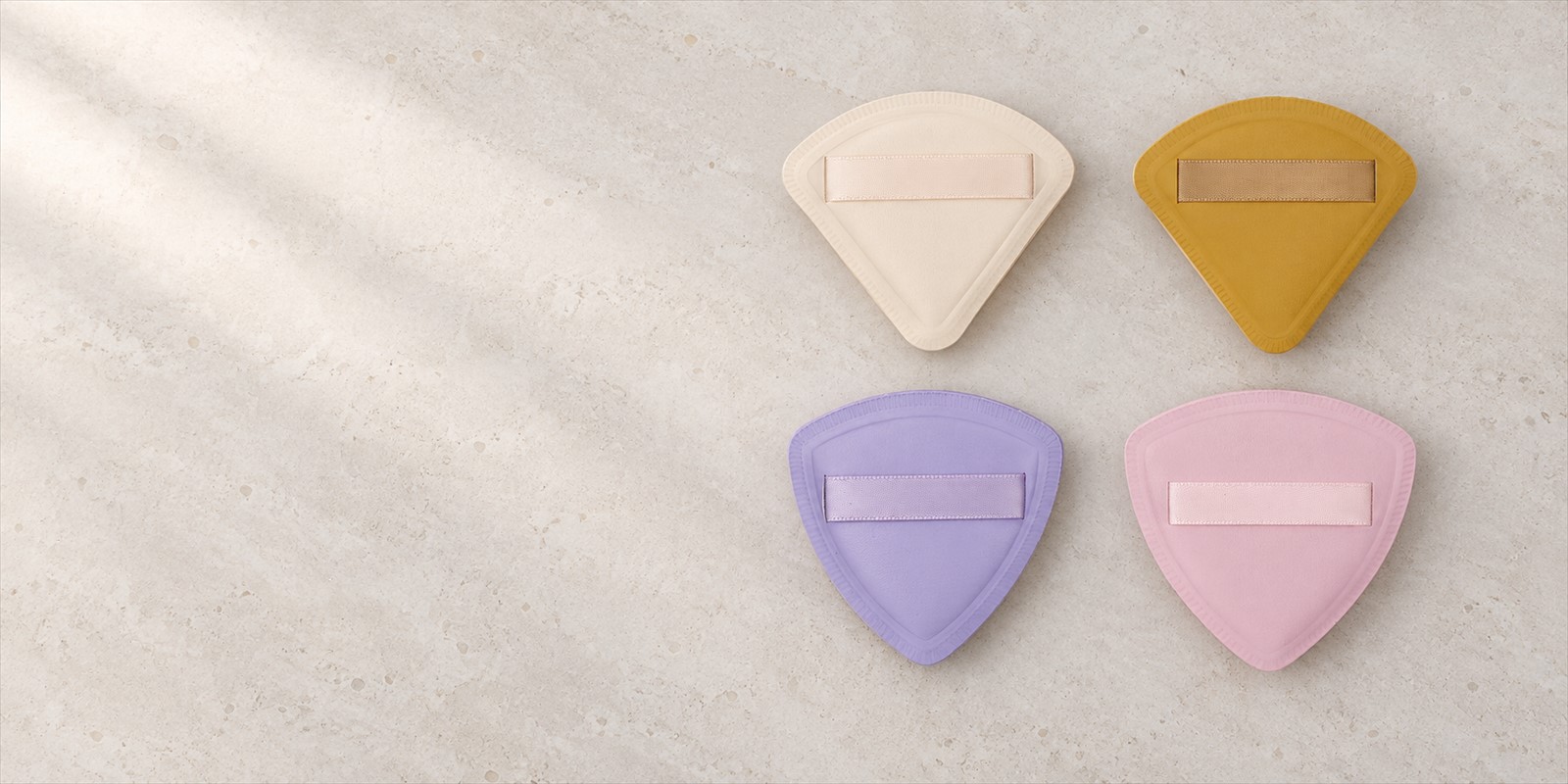 Four triangular makeup puffs in cream, mustard, lavender, and pink on a warm limestone surface